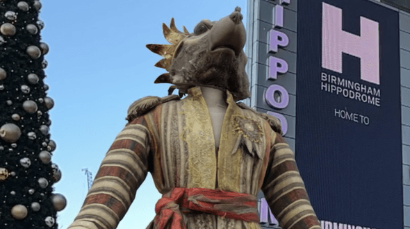 King Rat from The Nutcracker in augmented reality in front of Birmingham Hippodrome. King Rat has a detailed rat headpiece complete with crown, the jacket has stripped sleeves, shoulder pads, gold trim and a red band at the waist. The top half of the King Rat costume is standing in augmented reality to the left of a large, portrait screen attached to the front of Birmingham Hippodrome. To the left of King Rat is a Christmas tree with silver and gold baubles on. The sky in the background is blue, and there is a crane off in the distance.
