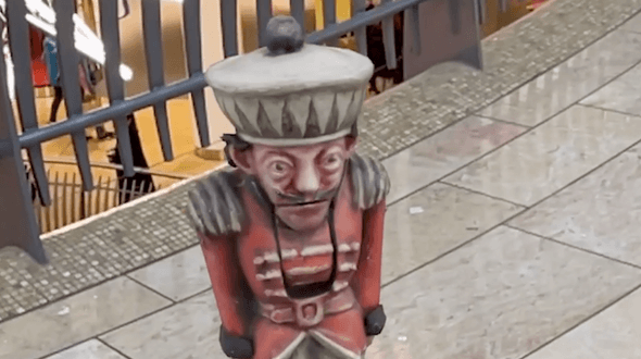 The Nutcracker doll from the ballet in The Bullring in augmented reality. The Nutcracker Doll has a red, painted jacket with shoulder detailing and a large, cream and black hat on top with a black sphere on top. The face of the doll has a neutral expression, and has a black moustache and black hair coming out of the back of the hat. The doll has its arms by its sides. From the waist down of the doll has been cropped from the image. The flooring in the Bullring is a cream marble effect. Behind the doll is a grey railing, behind which, you can see the downstairs level.