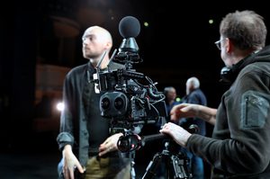 A complicated camera is the centre focus of the image, light shining off it’s matt black components. The camera is being held by a camera tripod and supported by Clive Booth’s left hand resting on the rig. The camera has various coiled wires coming out of it, antenna at the front, a dual lens and a spherical microphone sat on the top. Clive is facing away from the image, looking into the background. Clive has short brown hair, is wearing glasses and a black fleece. George Wood is standing in the background out of focus. George has shaved hair, a dark trimmed beard, is wearing beige trousers, a black t-shirt and grey over-shirt. There are two out of focus people in the background in conversation. A light is shining behind George and in the far background, balconies of the theatre can be seen. Image credit: Mark Spencer.