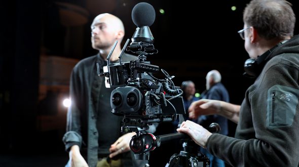 A complicated camera is the centre focus of the image, light shining off it’s matt black components. The camera is being held by a camera tripod and supported by Clive Booth’s left hand resting on the rig. The camera has various coiled wires coming out of it, antenna at the front, a dual lens and a spherical microphone sat on the top. Clive is facing away from the image, looking into the background. Clive has short brown hair, is wearing glasses and a black fleece. George Wood is standing in the background out of focus. George has shaved hair, a dark trimmed beard, is wearing beige trousers, a black t-shirt and grey over-shirt. There are two out of focus people in the background in conversation. A light is shining behind George and in the far background, balconies of the theatre can be seen. Image credit: Mark Spencer.