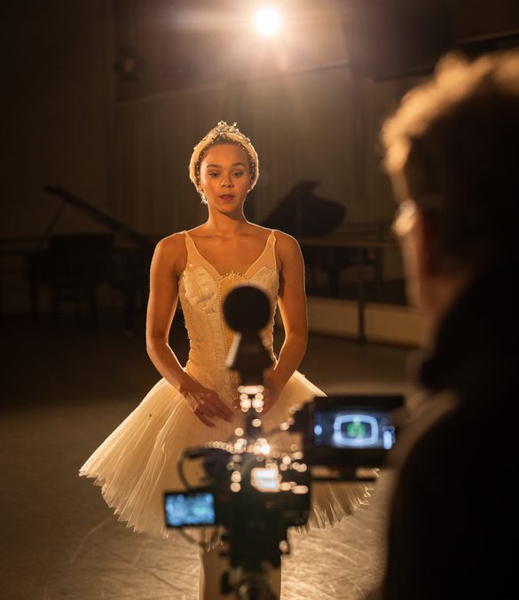 Regan Hutsell is standing in front of a virtual reality camera in a dimly lit studio and is the main focus of the image. Regan has dark hair tied back using a feathered headband with silver and pearl detailing. Regan is wearing a white tutu with pearl detailing from the ballet, Swan Lake. Clive Booth is standing to the right, but is out of camera focus. Clive has dark hair and is wearing glasses and a black top. The virtual reality camera that Clive is standing behind has multiple screens and a spherical microphone on the top. The camera is black and out of focus. A bright light is shining from behind Regan, and is lighting the studio as well as reflecting off of the camera and the sparkly detailing on Regan’s costume. In the background, there is a black, glossy grand piano, wall mounted TV, ballet barre and mirrors on the wall. The floor is grey with scuffs on it. Credit Mark Spencer.