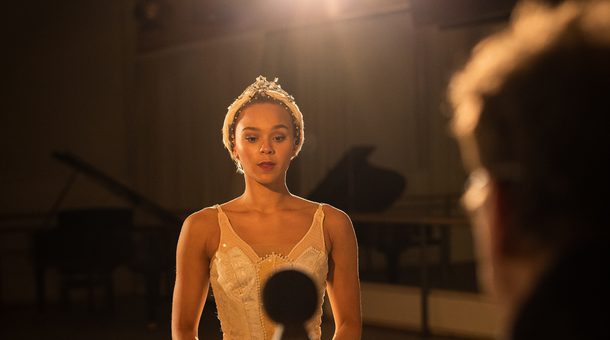 Regan Hutsell is standing in front of a virtual reality camera in a dimly lit studio and is the main focus of the image. Regan has dark hair tied back using a feathered headband with silver and pearl detailing. Regan is wearing a white tutu with pearl detailing from the ballet, Swan Lake. Clive Booth is standing to the right, but is out of camera focus. Clive has dark hair and is wearing glasses and a black top. The virtual reality camera that Clive is standing behind has multiple screens and a spherical microphone on the top. The camera is black and out of focus. A bright light is shining from behind Regan, and is lighting the studio as well as reflecting off of the camera and the sparkly detailing on Regan’s costume. In the background, there is a black, glossy grand piano, wall mounted TV, ballet barre and mirrors on the wall. The floor is grey with scuffs on it. Credit Mark Spencer.