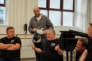 Josef Reed, Barry Kirby and Tom Bright of the Freefall Dance Company are sitting in a line from left to right of the image on chairs in a corner of a dance studio whilst George Wood from the filming team stands behind Barry whilst holding two white virtual reality (VR) headsets with both hands. George has shaved hair and a dark trimmed beard with a hooped earring in the left ear. George is wearing a white t-shirt, black trousers and a grey over-shirt. George is looking at Barry’s reaction to the headset content and is smiling. Clive Booth is kneeling down to the right of of Barry, looking at them and smiling as they react to the content. Clive has short brown hair and is wearing a light tan coloured jacket with red tartan lining. The Freefall Dancers are all wearing black t-shirts with the white Birmingham Royal Ballet text logo, below which is written ‘Freefall Dance’ also in white. Barry is smiling and looking to the right. Barry has very shaved dark hair and is sitting centrally in the image, raising both arms to chest height with both thumbs up. Josef is sitting on the left of the image and is looking at Barry. Josef has short brown hair and has their arms folded. Tom is sitting on the right side of the image and is also looking at Barry. Tom also has short brown hair but only a portion of Tom can be seen in the image as the frame cuts off half of their body. In the background of the image there is a black, glossy baby grand piano behind Clive and Barry. At the back of the image there are white panelled walls, large, red framed windows, a light brown ballet barre and to the right, a white curtain. Image credit: Andy Ford.