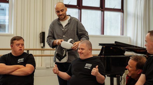Josef Reed, Barry Kirby and Tom Bright of the Freefall Dance Company are sitting in a line from left to right of the image on chairs in a corner of a dance studio whilst George Wood from the filming team stands behind Barry whilst holding two white virtual reality (VR) headsets with both hands. George has shaved hair and a dark trimmed beard with a hooped earring in the left ear. George is wearing a white t-shirt, black trousers and a grey over-shirt. George is looking at Barry’s reaction to the headset content and is smiling. Clive Booth is kneeling down to the right of of Barry, looking at them and smiling as they react to the content. Clive has short brown hair and is wearing a light tan coloured jacket with red tartan lining. The Freefall Dancers are all wearing black t-shirts with the white Birmingham Royal Ballet text logo, below which is written ‘Freefall Dance’ also in white. Barry is smiling and looking to the right. Barry has very shaved dark hair and is sitting centrally in the image, raising both arms to chest height with both thumbs up. Josef is sitting on the left of the image and is looking at Barry. Josef has short brown hair and has their arms folded. Tom is sitting on the right side of the image and is also looking at Barry. Tom also has short brown hair but only a portion of Tom can be seen in the image as the frame cuts off half of their body. In the background of the image there is a black, glossy baby grand piano behind Clive and Barry. At the back of the image there are white panelled walls, large, red framed windows, a light brown ballet barre and to the right, a white curtain. Image credit: Andy Ford.