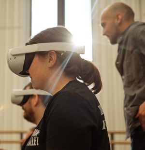 To the left of the photo, two people are can be seen from the chest up, one in front of the other, looking to the left. They are wearing black teeshirts with indistinguishable white logos and white VR headsets. The person in front is in sharp focus; she has her brown hair in a ponytail. The person behind is in soft focus; he has a beard. They are members of the Freefall Dance Company. They are sitting in a light-filled ballet studio, with a window and the ballet barre visible behind them. Light is pouring in through the window creating rays that are reflecting off the person at the front’s headset. George Wood from Canon is out of camera focus, standing behind them, wearing a grey overshirt and jeans. George has a shaven head and trimmed dark stubble. The studio walls are white, and there is a wooden ballet barre on the back wall. Image Credit: Andy Ford.