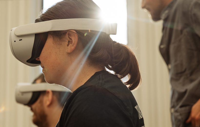 To the left of the photo, two people are can be seen from the chest up, one in front of the other, looking to the left. They are wearing black teeshirts with indistinguishable white logos and white VR headsets. The person in front is in sharp focus; she has her brown hair in a ponytail. The person behind is in soft focus; he has a beard. They are members of the Freefall Dance Company. They are sitting in a light-filled ballet studio, with a window and the ballet barre visible behind them. Light is pouring in through the window creating rays that are reflecting off the person at the front’s headset. George Wood from Canon is out of camera focus, standing behind them, wearing a grey overshirt and jeans. George has a shaven head and trimmed dark stubble. The studio walls are white, and there is a wooden ballet barre on the back wall. Image Credit: Andy Ford.