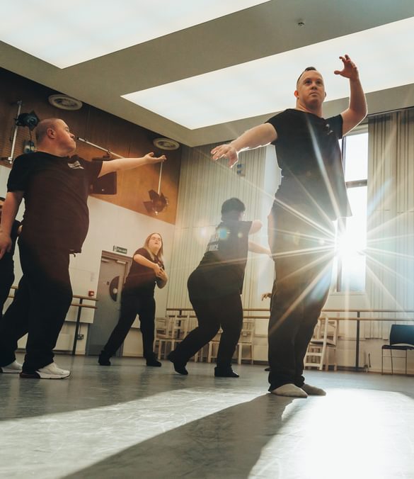 Six members of the Freefall Dance Company are performing in a dance studio. The performers, wearing black t-shirts with a white Birmingham Royal Ballet logo on the front left, and a larger white logo at the rear, with ‘Freefall Dance Company’ on are biased towards the left of the image in various poses.  The dancers are lit by the sun streaming in from the two large dark red framed windows on the rear wall. The foremost performer is standing with their left arm raised, and right arm pointing towards the floor whilst looking off into the distance as the sun shines from behind them, their shadow is being cast onto the floor. Towards the back of the room there is a ballet barre, a glossy black baby grand piano, and some stacked black chairs. The studio has large rectangular light panels in the ceiling, with half wooden panelled and half white walls. The floor is a matt grey with scuffs on it. Credit Andy Ford.