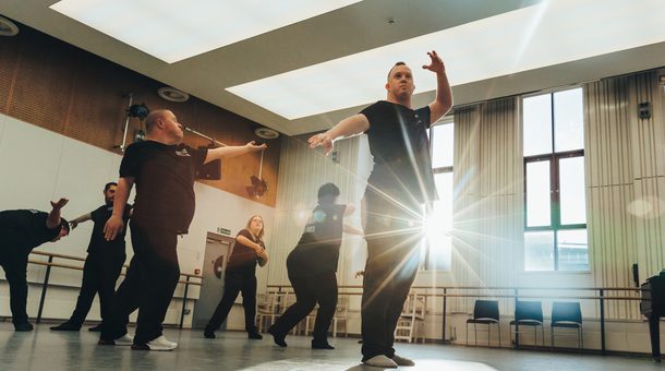 Six members of the Freefall Dance Company are performing in a dance studio. The performers, wearing black t-shirts with a white Birmingham Royal Ballet logo on the front left, and a larger white logo at the rear, with ‘Freefall Dance Company’ on are biased towards the left of the image in various poses. The dancers are lit by the sun streaming in from the two large dark red framed windows on the rear wall. The foremost performer is standing with their left arm raised, and right arm pointing towards the floor whilst looking off into the distance as the sun shines from behind them, their shadow is being cast onto the floor. Towards the back of the room there is a ballet barre, a glossy black baby grand piano, and some stacked black chairs. The studio has large rectangular light panels in the ceiling, with half wooden panelled and half white walls. The floor is a matt grey with scuffs on it. Credit Andy Ford.