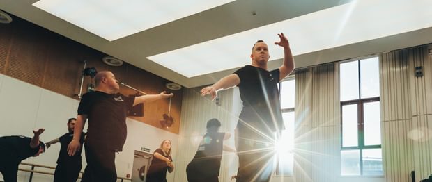 Six members of the Freefall Dance Company are performing in a dance studio. The performers, wearing black t-shirts with a white Birmingham Royal Ballet logo on the front left, and a larger white logo at the rear, with ‘Freefall Dance Company’ on are biased towards the left of the image in various poses.  The dancers are lit by the sun streaming in from the two large dark red framed windows on the rear wall. The foremost performer is standing with their left arm raised, and right arm pointing towards the floor whilst looking off into the distance as the sun shines from behind them, their shadow is being cast onto the floor. Towards the back of the room there is a ballet barre, a glossy black baby grand piano, and some stacked black chairs. The studio has large rectangular light panels in the ceiling, with half wooden panelled and half white walls. The floor is a matt grey with scuffs on it. Credit Andy Ford.