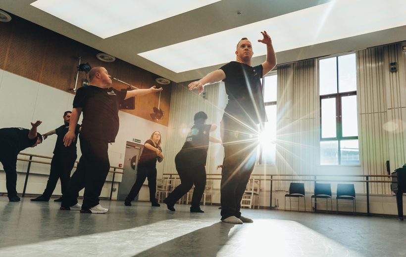 Six members of the Freefall Dance Company are performing in a dance studio. The performers, wearing black t-shirts with a white Birmingham Royal Ballet logo on the front left, and a larger white logo at the rear, with ‘Freefall Dance Company’ on are biased towards the left of the image in various poses. The dancers are lit by the sun streaming in from the two large dark red framed windows on the rear wall. The foremost performer is standing with their left arm raised, and right arm pointing towards the floor whilst looking off into the distance as the sun shines from behind them, their shadow is being cast onto the floor. Towards the back of the room there is a ballet barre, a glossy black baby grand piano, and some stacked black chairs. The studio has large rectangular light panels in the ceiling, with half wooden panelled and half white walls. The floor is a matt grey with scuffs on it. Credit Andy Ford.