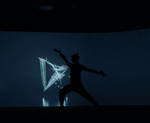 A dancer in shadow is stood side-on to the camera with left arm raised and right arm pointing towards the right side of the image. The dancer is wearing a baseball cap and has motion sensors on each wrist, ankle and on the hat itself. Behind the dancer there is a large screen which is displaying shades of blue and digital triangles of blue light which are interacting with the dancer’s movements.