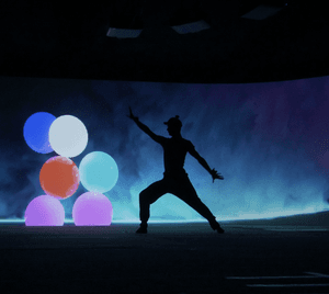 A shadow of a dancer is standing out against a smokey blue background with multi-colour balloons. The dancer is completely in shadow, with the left arm raised and right arm out pointing towards the floor, their left leg is bent at the knee with their weight leaning towards it, the right leg is straight and pointed to the right. In the background there is a smokey blue haze on a screen, with hints of purple. In the centre of the screen there are six balloons in red, blue, white, light blue and purple. In the ceiling two white projectors are mounted in an arch.