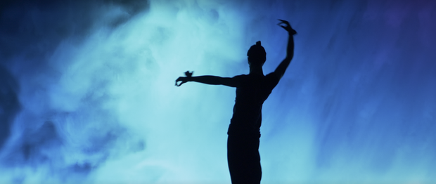 A shadow of a dancer is standing out against a smokey blue background. The dancer is completely in shadow, with the right arm raised in a slight arch, and left arm out level. The dancer’s body is slightly curved. The blue background is a large screen displaying smoke in hues of blue.