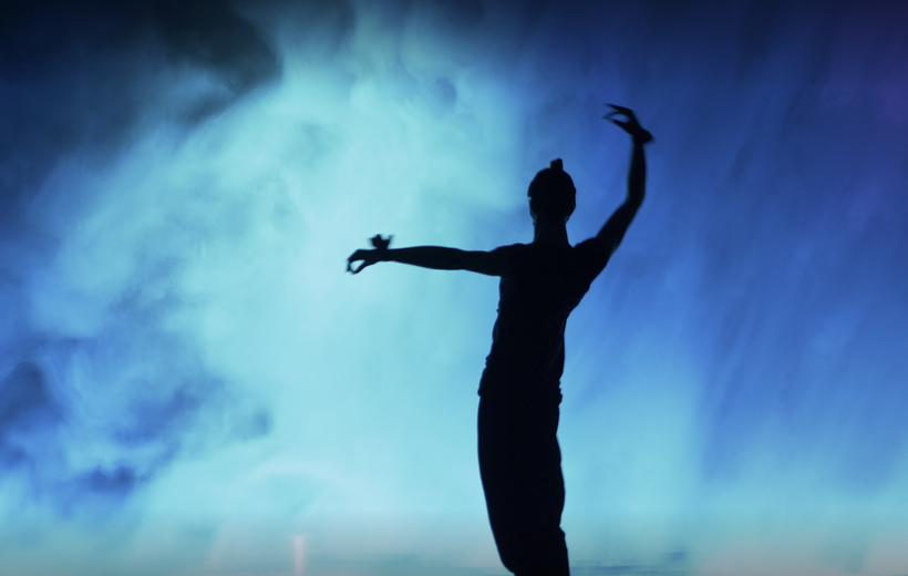 A shadow of a dancer is standing out against a smokey blue background. The dancer is completely in shadow, with the right arm raised in a slight arch, and left arm out level. The dancer’s body is slightly curved. The blue background is a large screen displaying smoke in hues of blue.