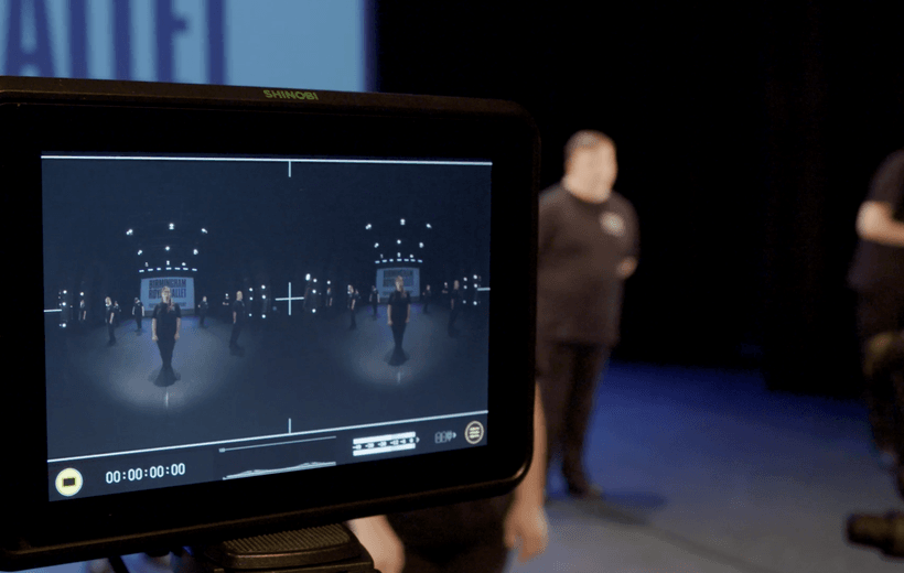 There is a large ‘SHINOBI’ screen on the left side of the image which is attached on a tripod. On the screen members of the Freefall Dance Company can be seen standing in a circular formation, wearing black t-shirts with the Freefall Dance Company logo in white on the left of the shirts. Behind the dancers on the screen is a large screen with a white background and black text which reads ‘Birmingham Royal Ballet, Freefall Dance Company.’ Stage lights can be seen above and to thew side of the dancers. At the bottom of the screen there is a timestamp and various tech icons in yellow. To the right of the screen there are two Freefall dancers that are out of camera focus. Behind the screen is the large white screen, but only the word ‘Ballet’ can be seen.