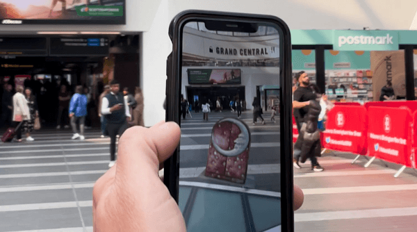 The image is of a hand holding a phone up to the main concourse in New Street station, which is dotted with members of the public. On the phone you can see the AR projection of Harlequin's box, a prop from The Nutcracker, apparently placed into the station space.