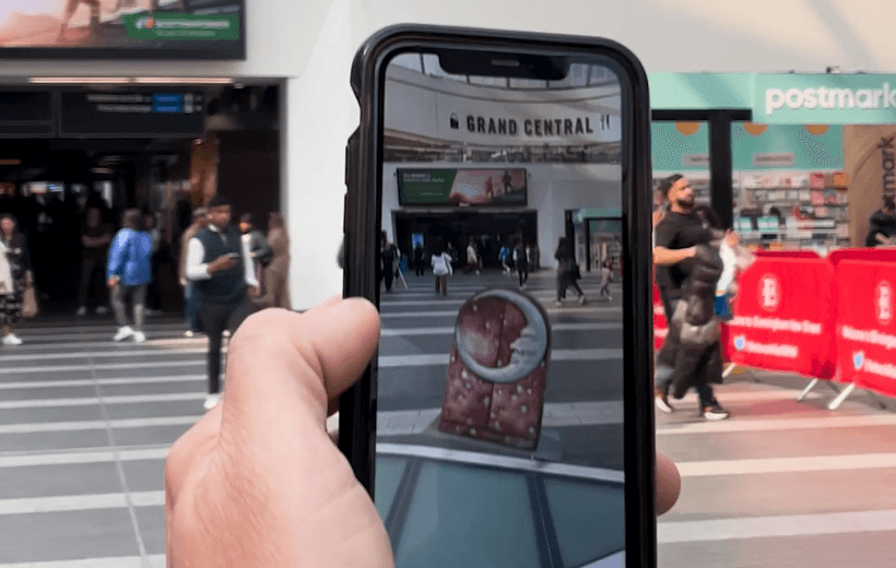 The image is of a hand holding a phone up to the main concourse in New Street station, which is dotted with members of the public. On the phone you can see the AR projection of Harlequin's box, a prop from The Nutcracker, apparently placed into the station space.